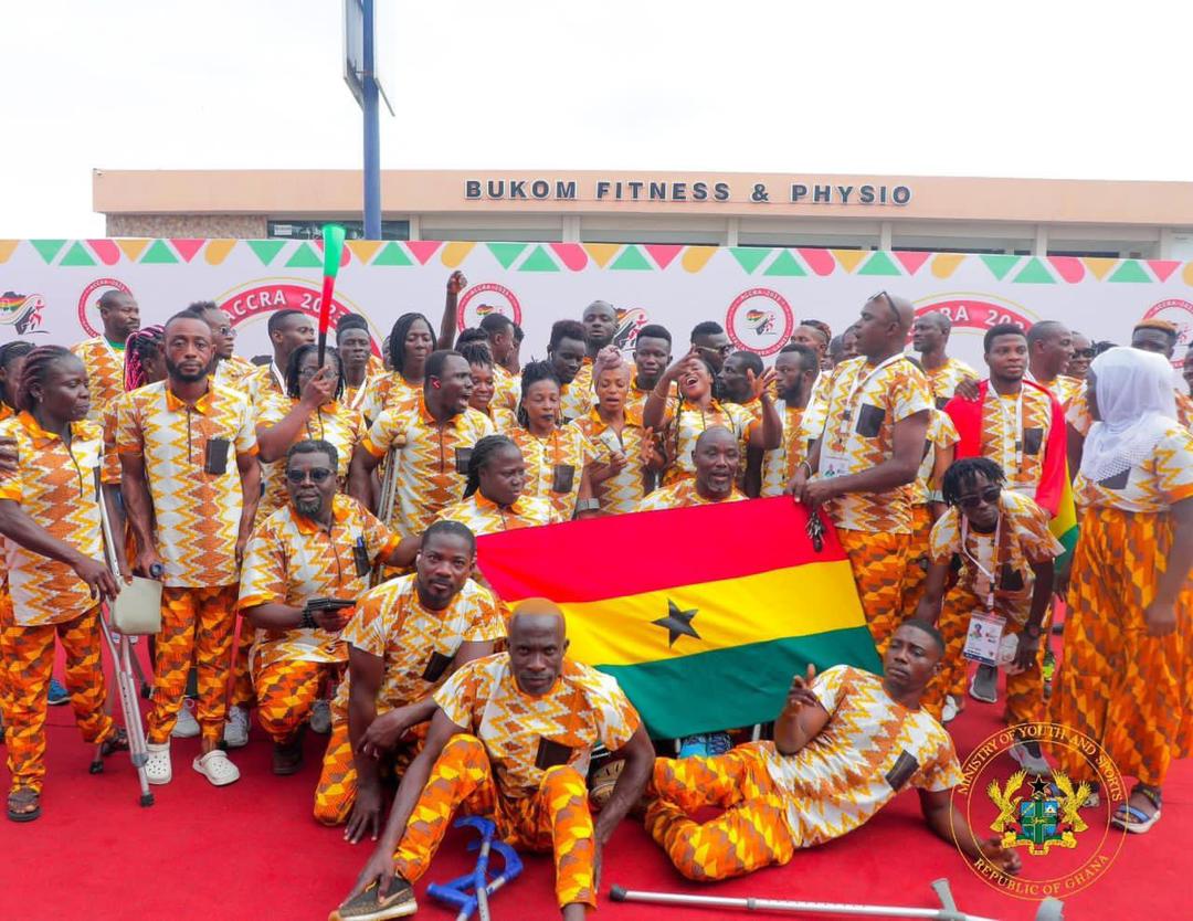Les membres de la délégation ghanéenne, vêtus de tenues assorties à motifs, se rassemblent autour du drapeau du Ghana ; plusieurs athlètes utilisent des béquilles.