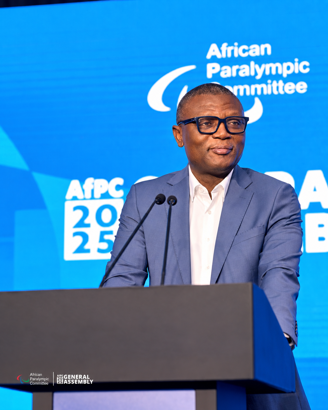 A speaker wearing glasses and a grey suit at the lectern against the blue AfPC General Assembly backdrop.