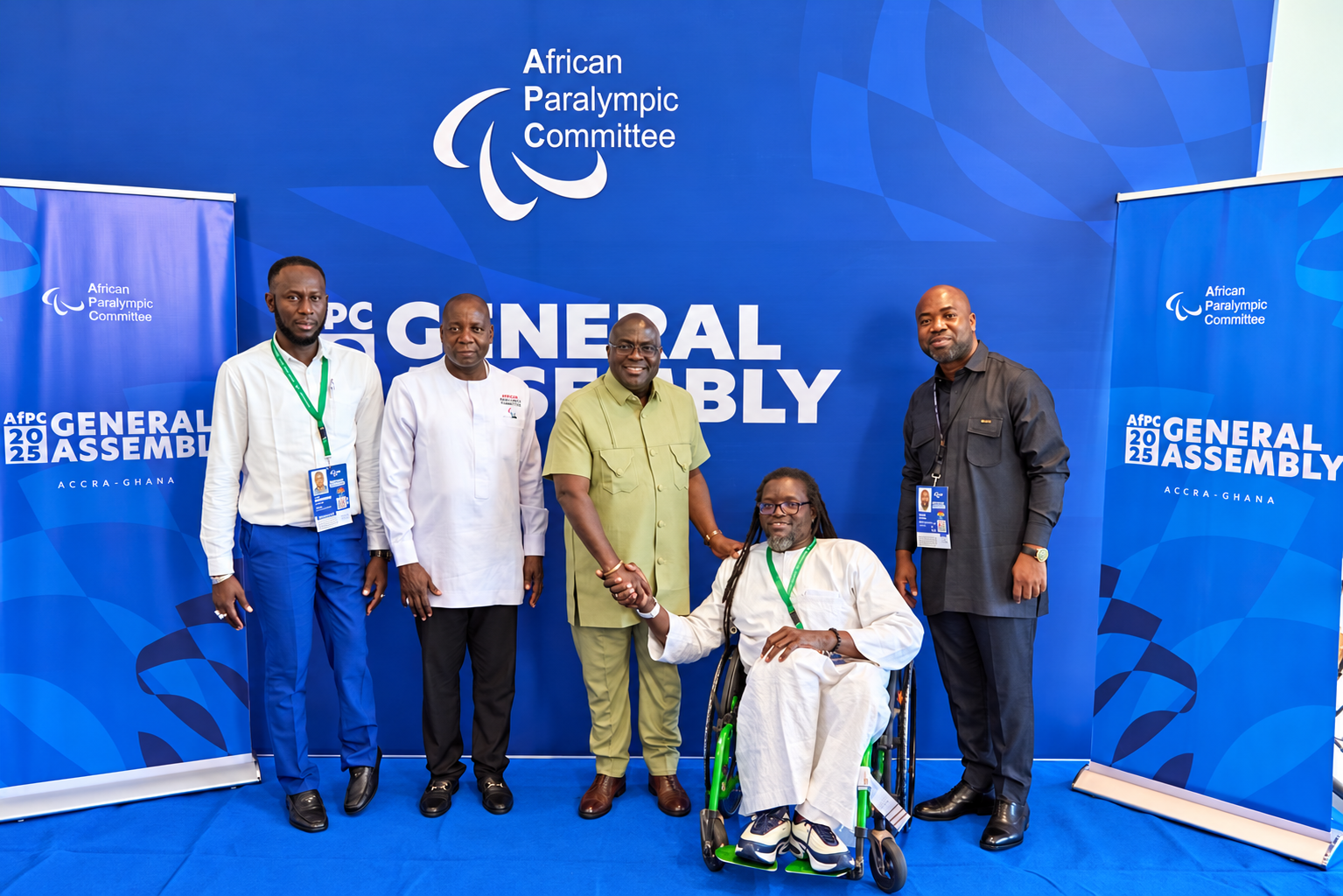 Five participants pose together, with a handshake beside a wheelchair in the foreground.