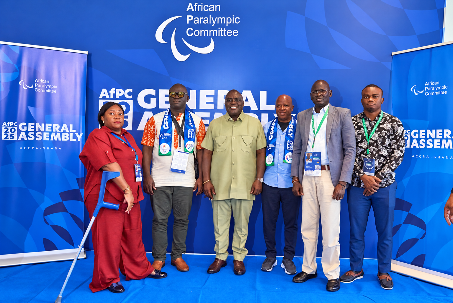 Six participants pose at the Assembly backdrop, including a woman using a blue forearm crutch.